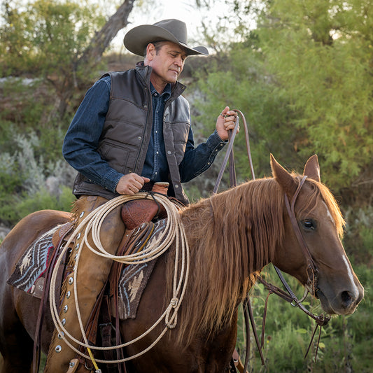 Stran Smith wearing a brown puffer vest with snap and zip front closure and leather detailing, layered over a denim long sleeve. He is holding a rope while riding a brown horse in a field.