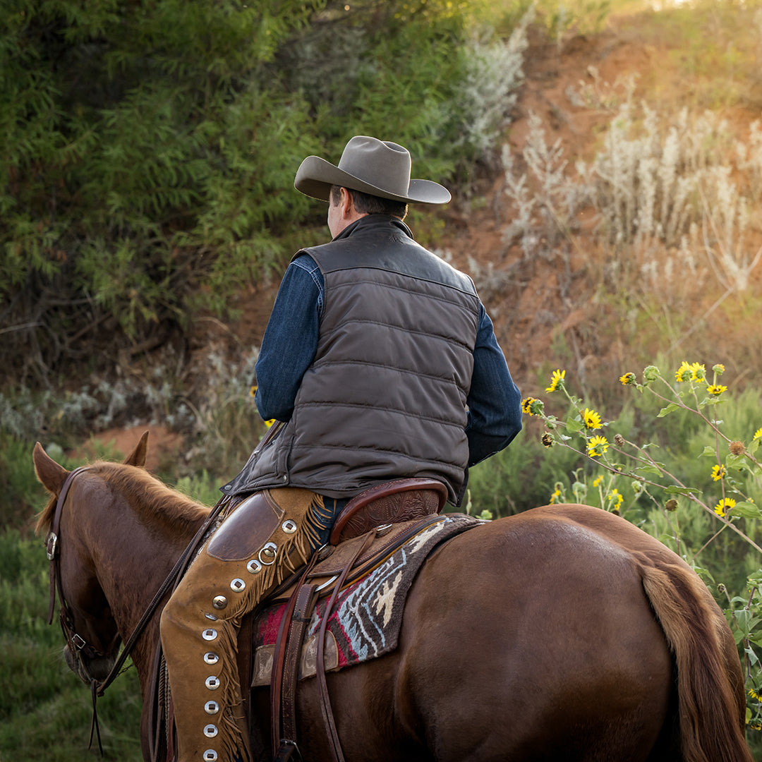 the back of Stran Smith wearing a brown puffer vest with leather detailing, layered over a denim long sleeve. He is holding a rope while riding a brown horse in a field.