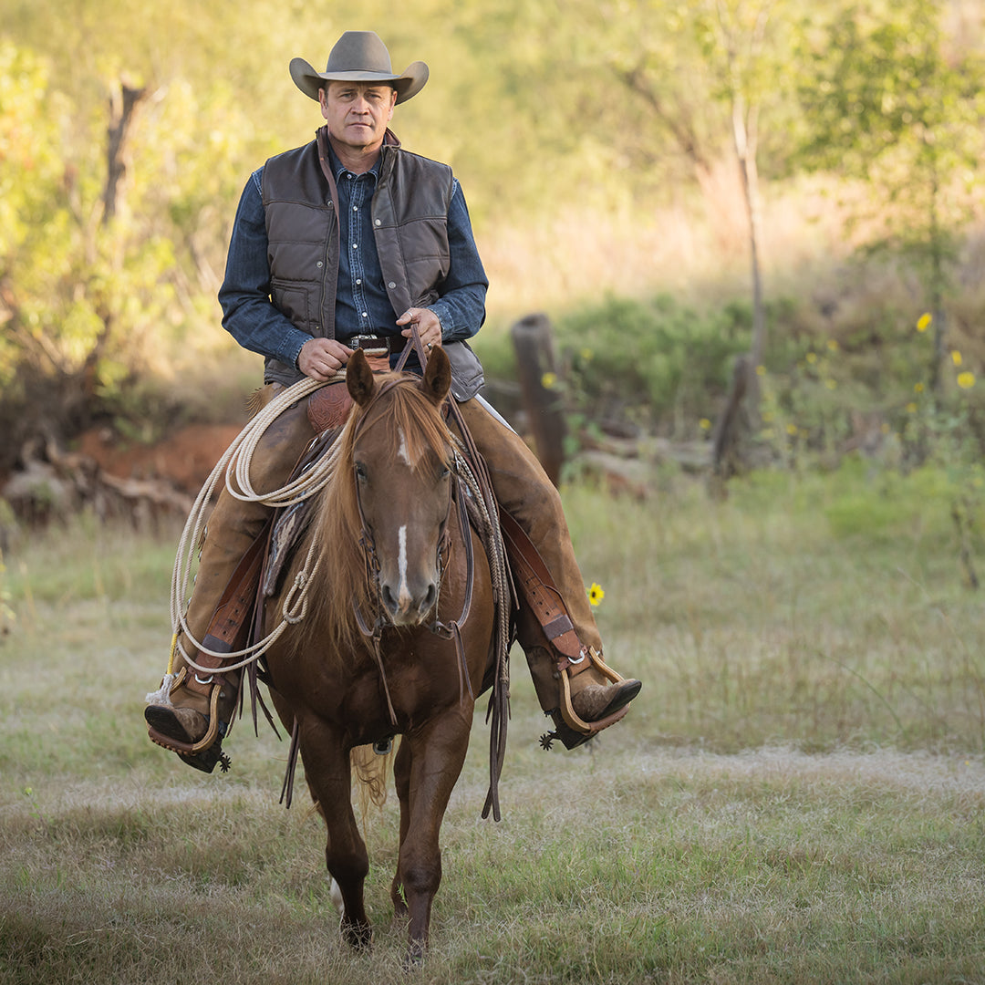 Stran Smith wearing a brown puffer vest with snap and zip front closure and leather detailing, layered over a denim long sleeve. He is holding a rope while riding a brown horse in a field.