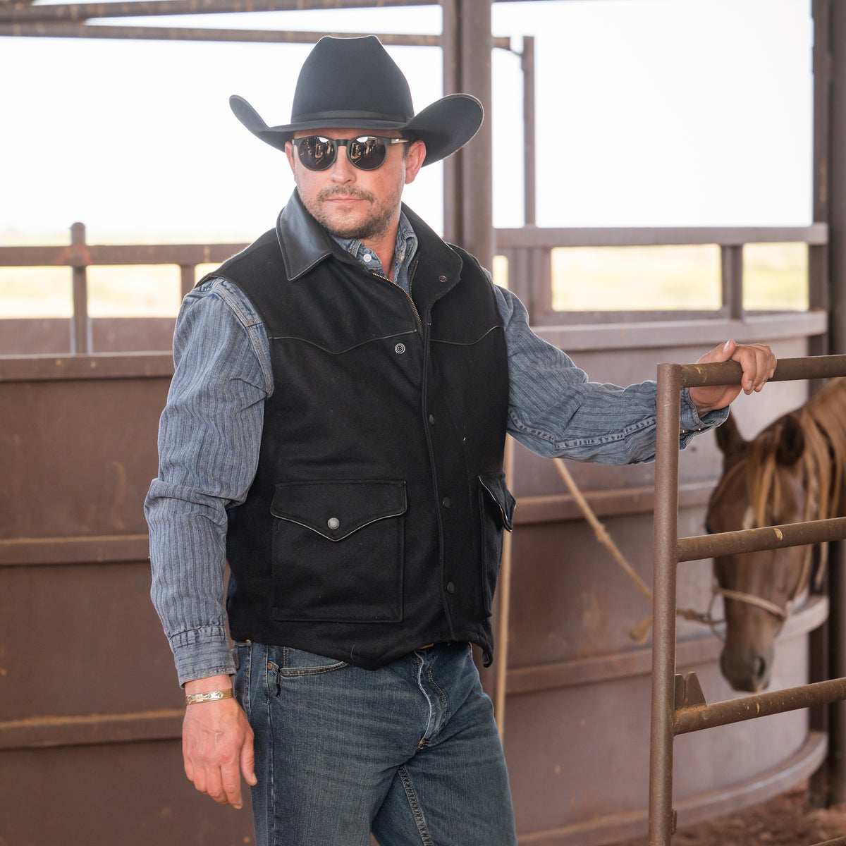 Man wearing sunglasses, a cowboy hat and our black wool vest with leather collar front zip and snap closure and two waist pockets in front of a brown horse