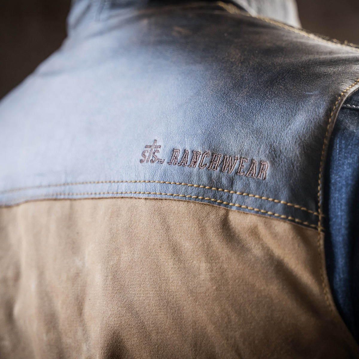 closeup of STS Ranchwear embroidered on the back of our snap and zip closure cotton canvas vest with full grain leather accents