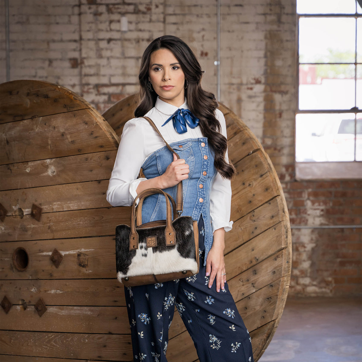 Soni Vest is wearing a multitextured and layered, white and blue outfit. She is standing in front of a large wooden barrel wearing a cowhide and leather purse using it's adjustable crossbody strap