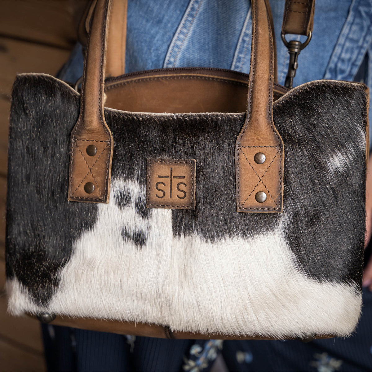 Close up of a cowhide and leather purse with two carry handles, removable adjustable crossbody strap and a center zip closure in front of denim 