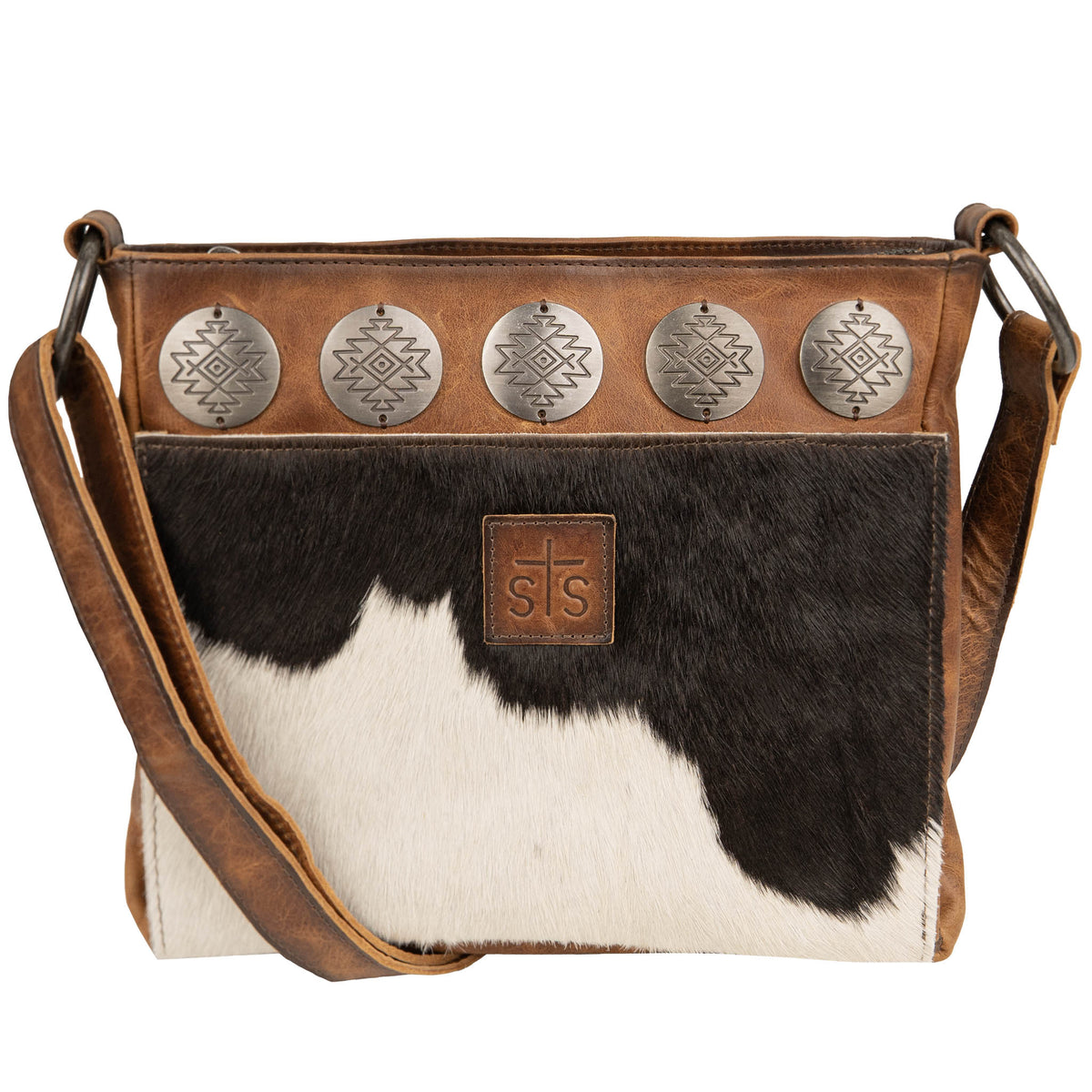 A brown and white cowhide crossbody bag with genuine leather accents, rustic metal conchos, an adjustable leather shoulder strap, and a zip top closure.