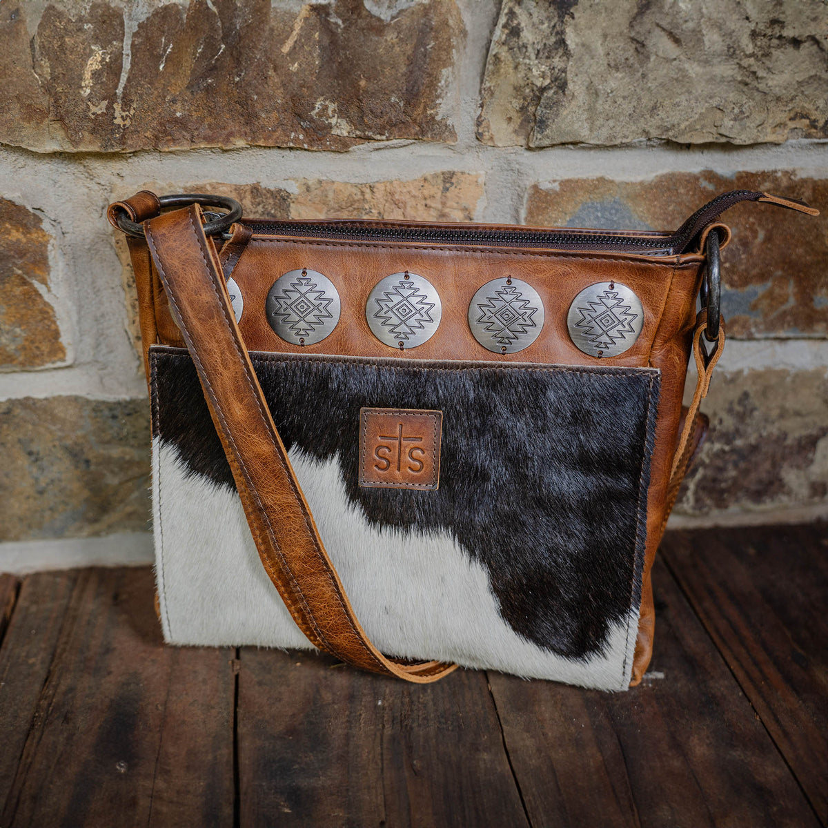 A brown and white cowhide crossbody bag with genuine leather accents, rustic metal conchos, an adjustable leather shoulder strap, and a zip top closure.