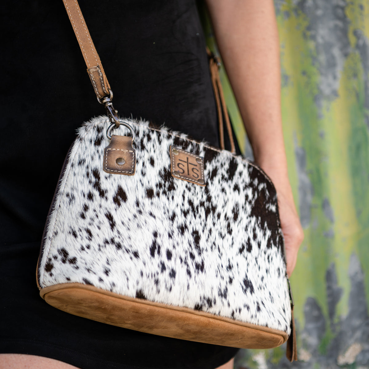 Person wearing a black dress and a white cowhide hair on hide leather pouch with brown genuine leather strap, zip around closure and leather patch with embossed STS logo.