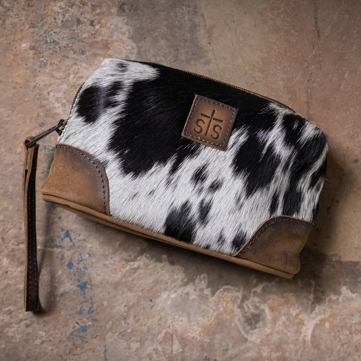 A small zippered cosmetic pouch with a black and white cowhide pattern, brown leather trim, and a brown leather patch featuring embossed STS logo.