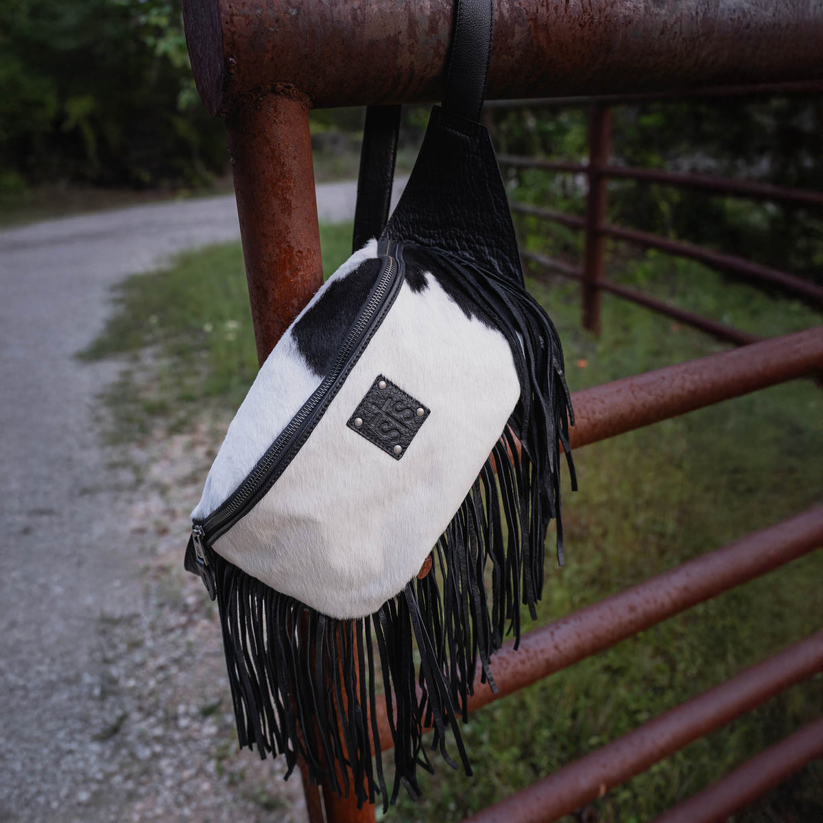 White crossbody bag with black fringe and leather patch hanging on a rusty metal fence.