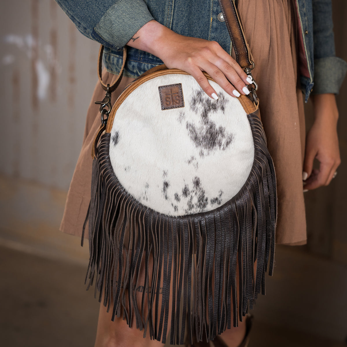 closeup of Style Brazile's hand holding our circle cowhide and distressed full grain leather purse with top zip closure, top zip closure and fringe along the bottom with an adjustable crossbody strap in front of wooden barn wall
