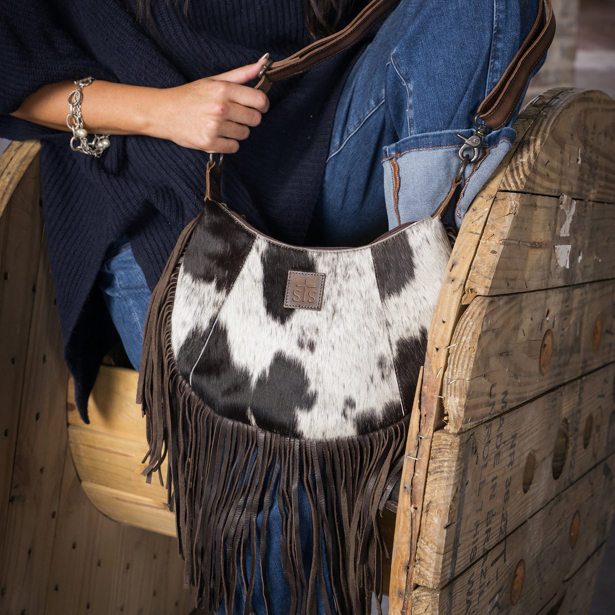 Whitney Vest holding our leather and cowhide crescent bag with fringe along the bottom on wooden barrel wheel