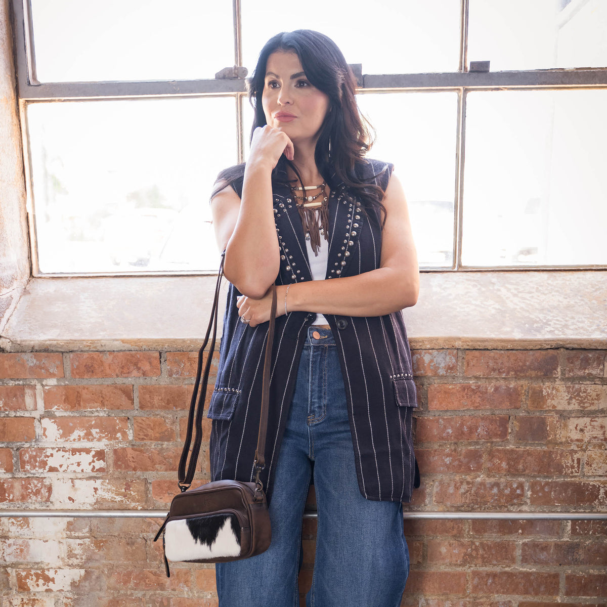 Whitney Vest wearing a cowhide and STS embossed leather crossbody purse leaning against a brick wall and windows