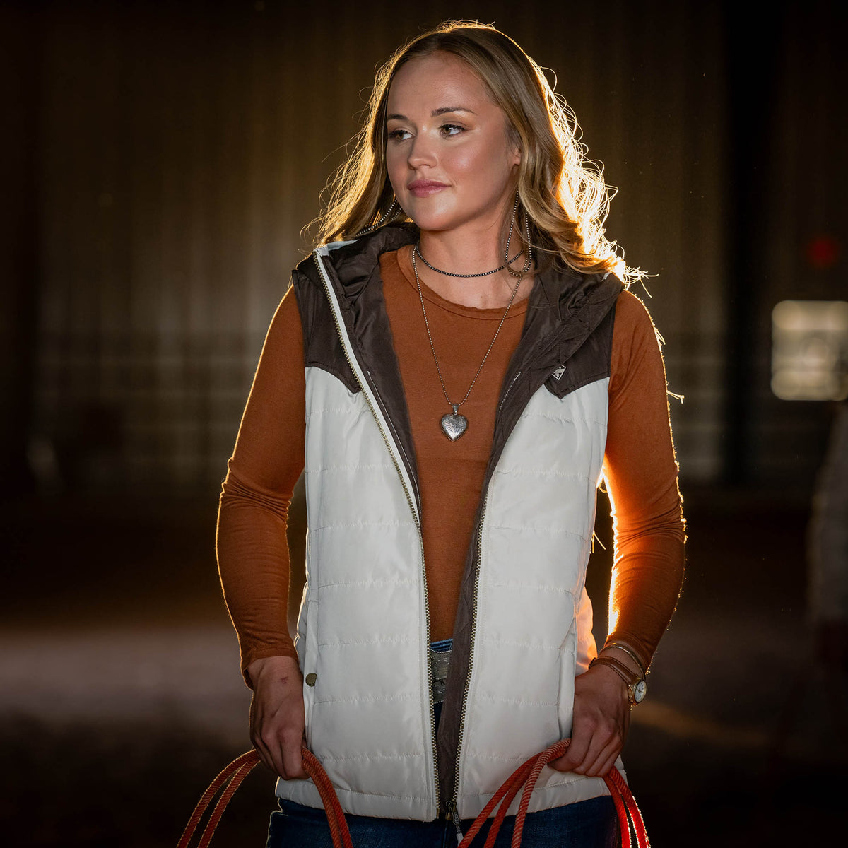 Woman wearing an orange long sleeve shirt and beige puffy vest holding a rope in a dimly lit indoor barn.