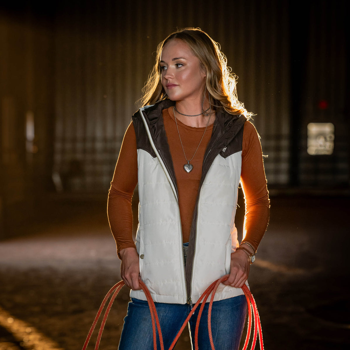 Woman wearing an orange long sleeve shirt and beige puffy vest holding a rope in a dimly lit indoor barn.