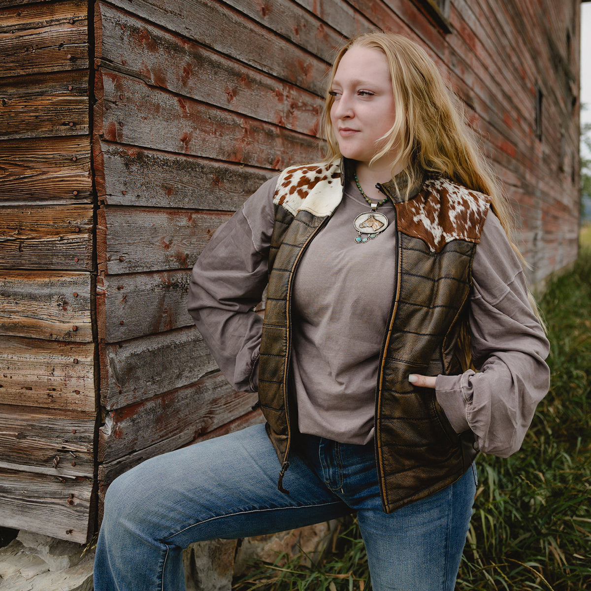 A woman wearing a brown quilted vest with cow-print faux fur trim over a taupe long-sleeve shirt and blue jeans, posing outdoors against a rustic wooden wall.