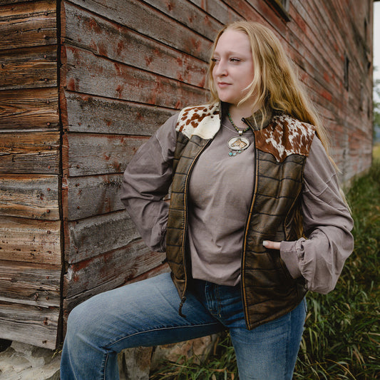 A woman wearing a brown quilted vest with cow-print faux fur trim over a taupe long-sleeve shirt and blue jeans, posing outdoors against a rustic wooden wall.
