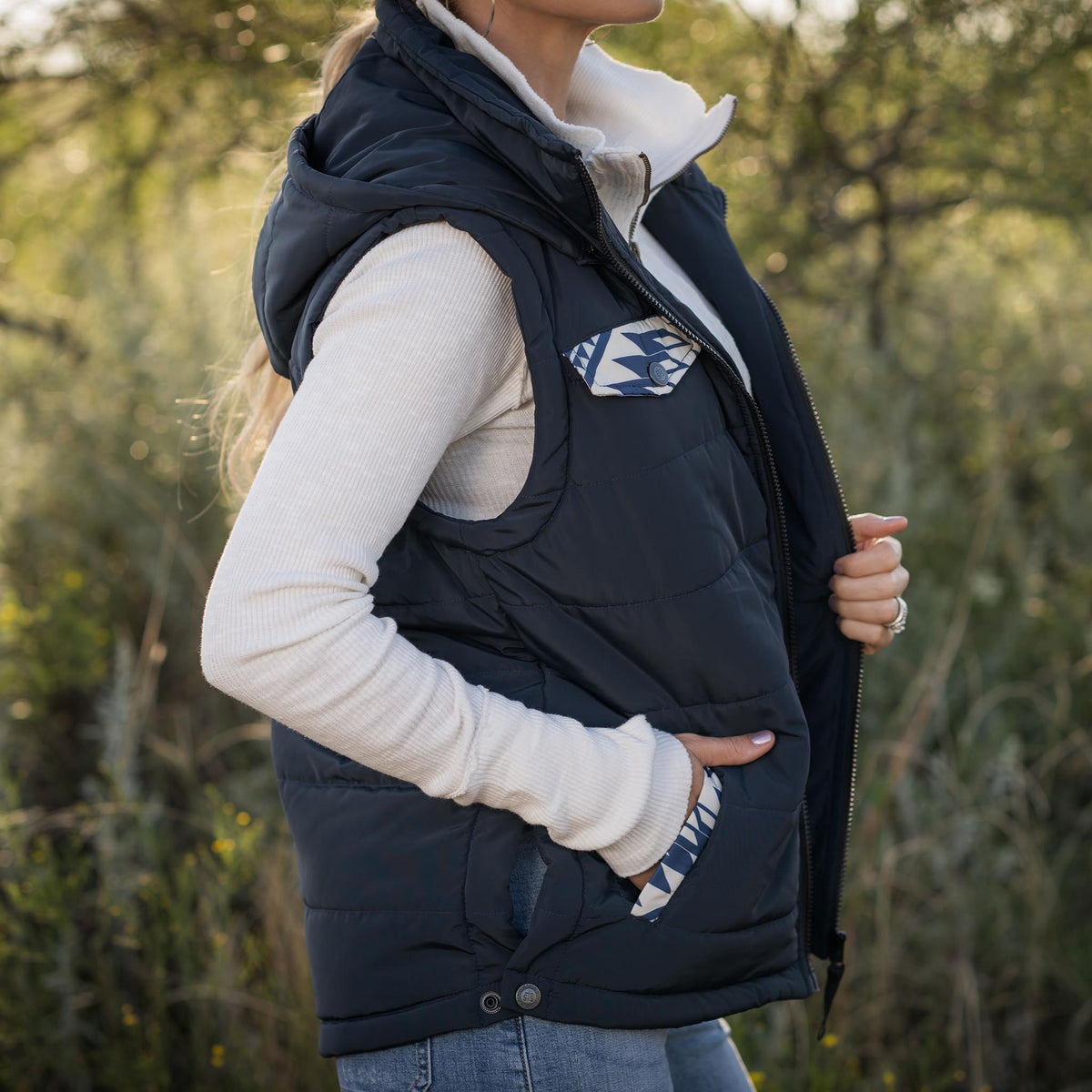 Person in a forest setting wearing a white long sleeve shirt, jeans and navy puffer style women's vest with zip front closure, hood and decorative aztec accents on chest and hand pockets.