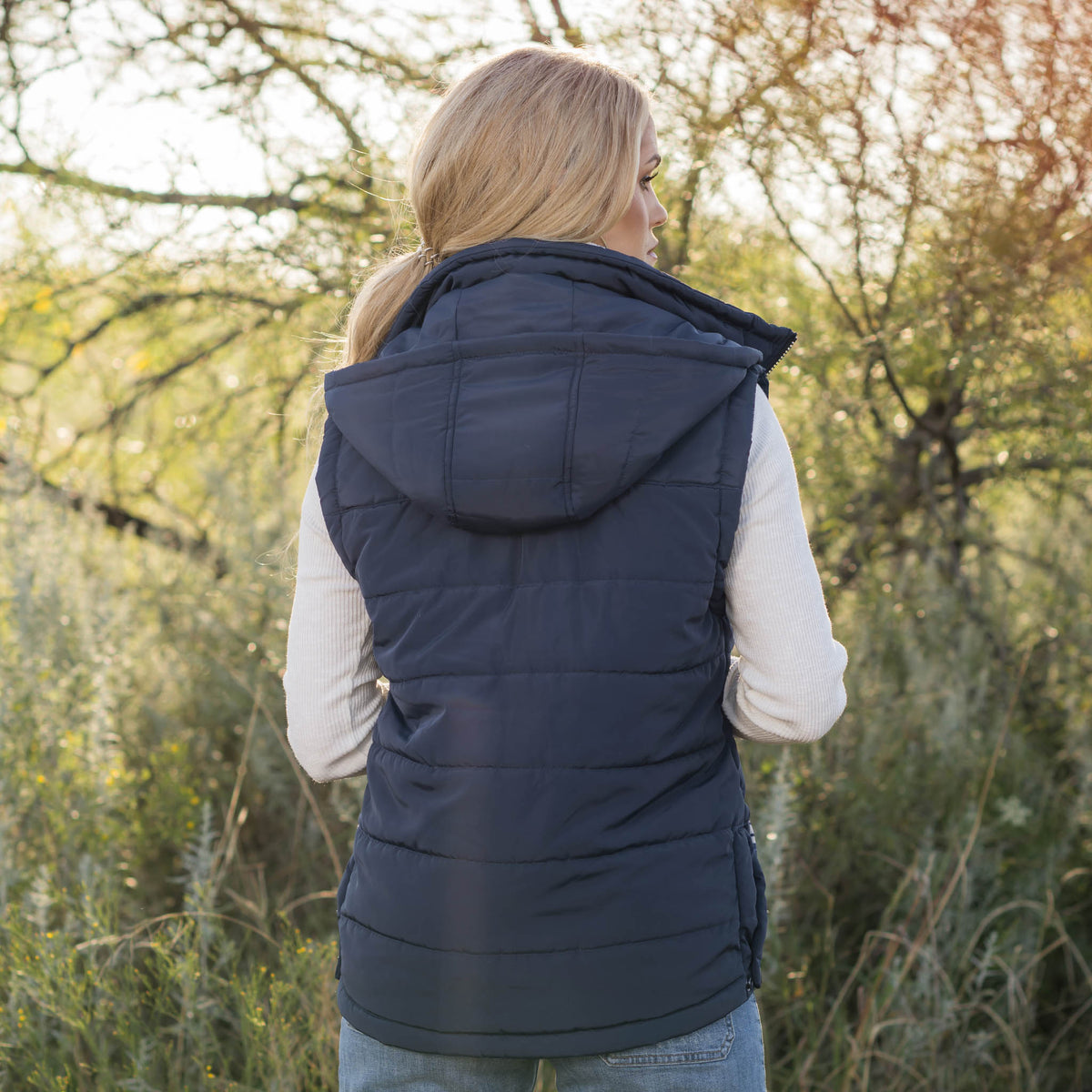 Person in a forest setting wearing a white long sleeve shirt, jeans and navy puffer style women's vest with a hood.
