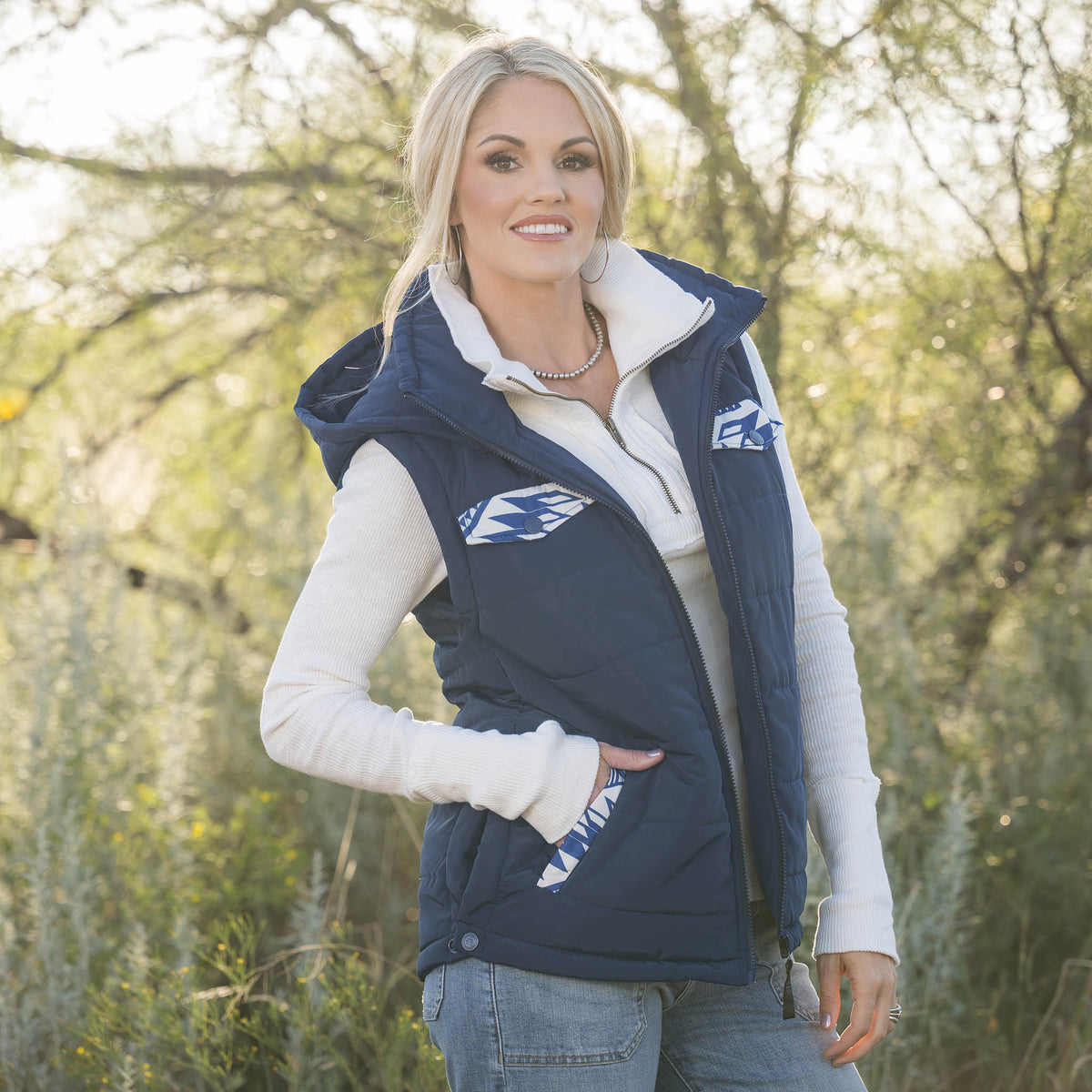 Woman in a forest setting wearing a white long sleeve shirt, jeans and navy puffer style women's vest with zip front closure, hood and decorative aztec accents on chest and hand pockets.