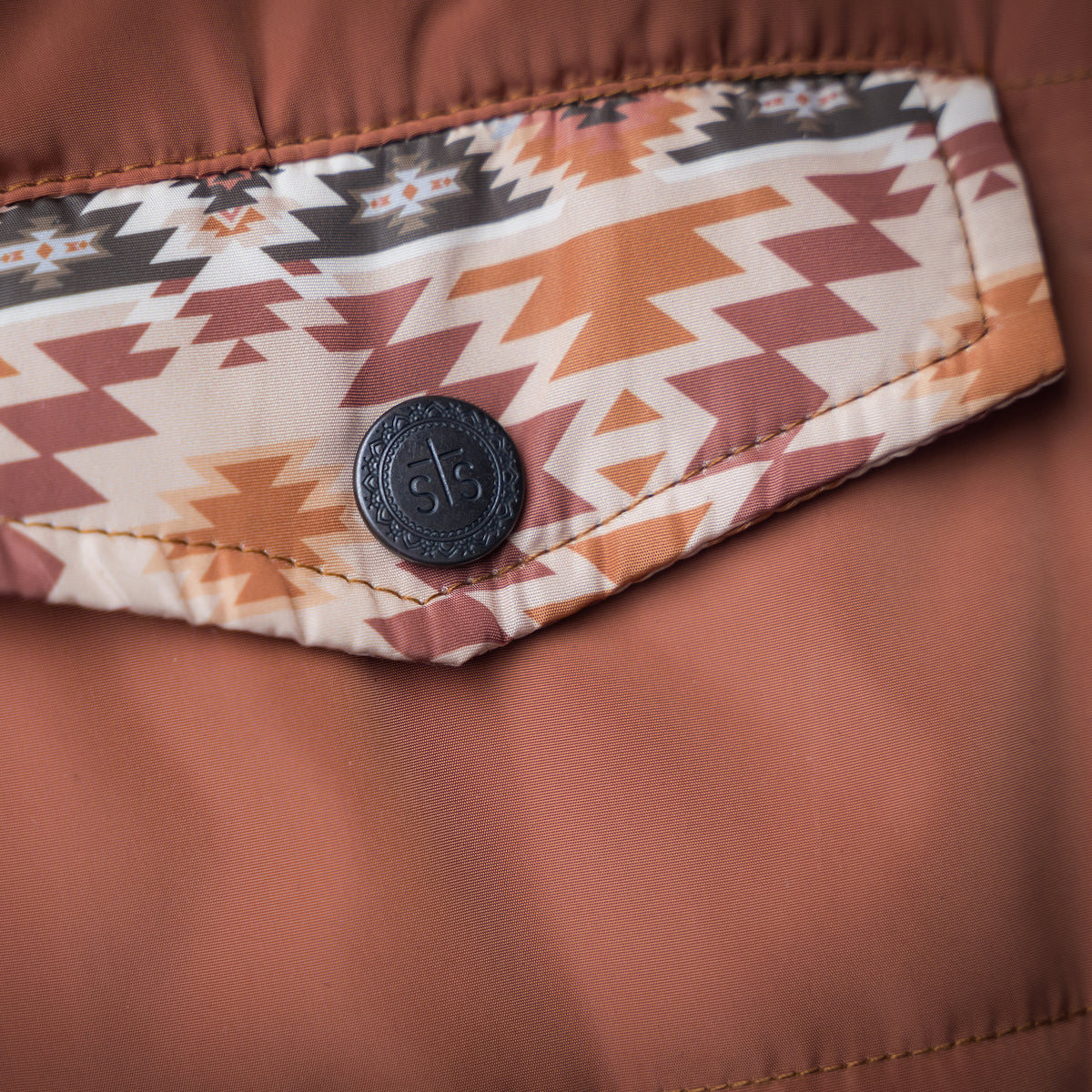 Closeup of the chest pocket of a rust orange zip up puffer vest with aztec accents.