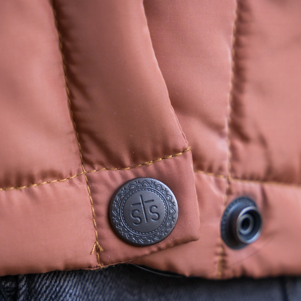 Closeup of the bottom snap of our rust orange puffer vest with STS stamped on the button.