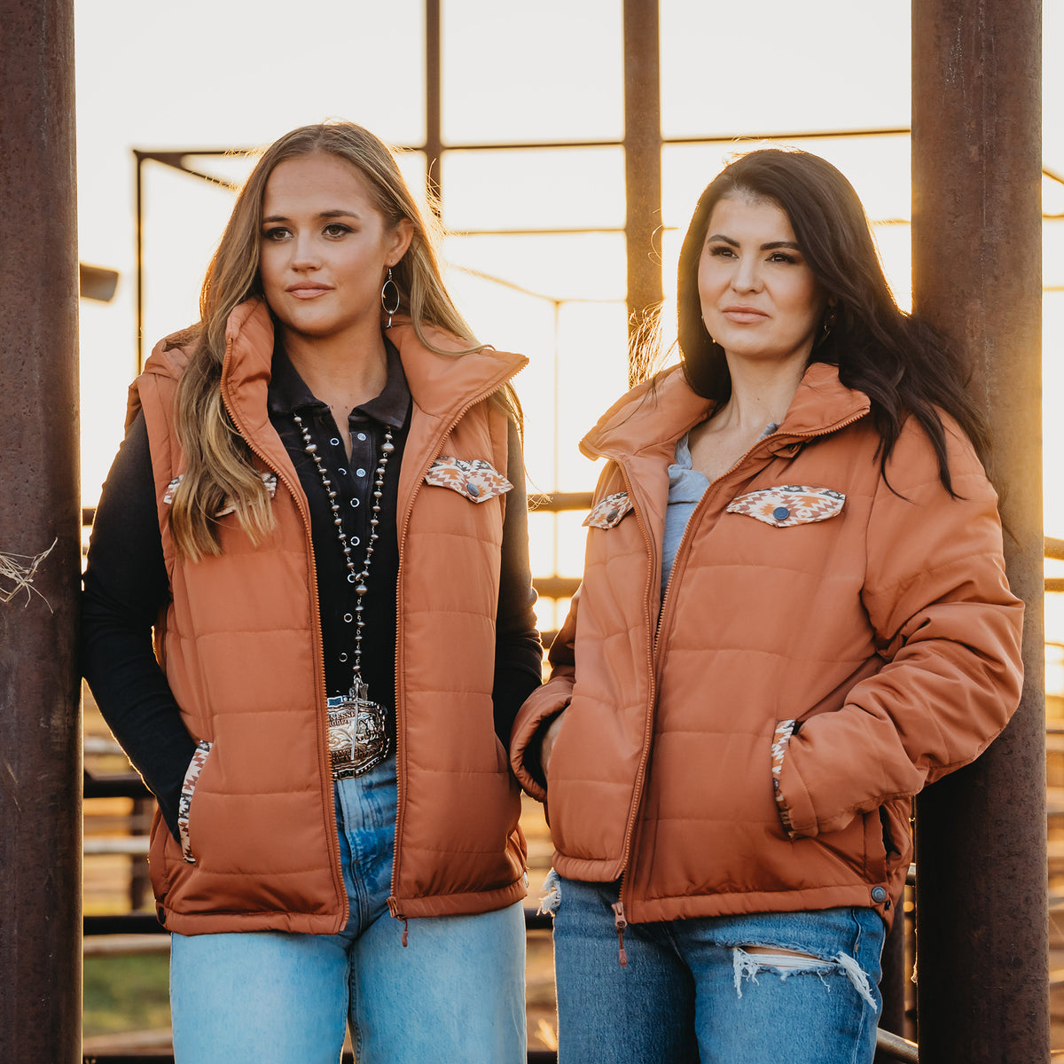Two women standing outside both wearing a rust orange zip up puffer vest with aztec accents and jeans.
