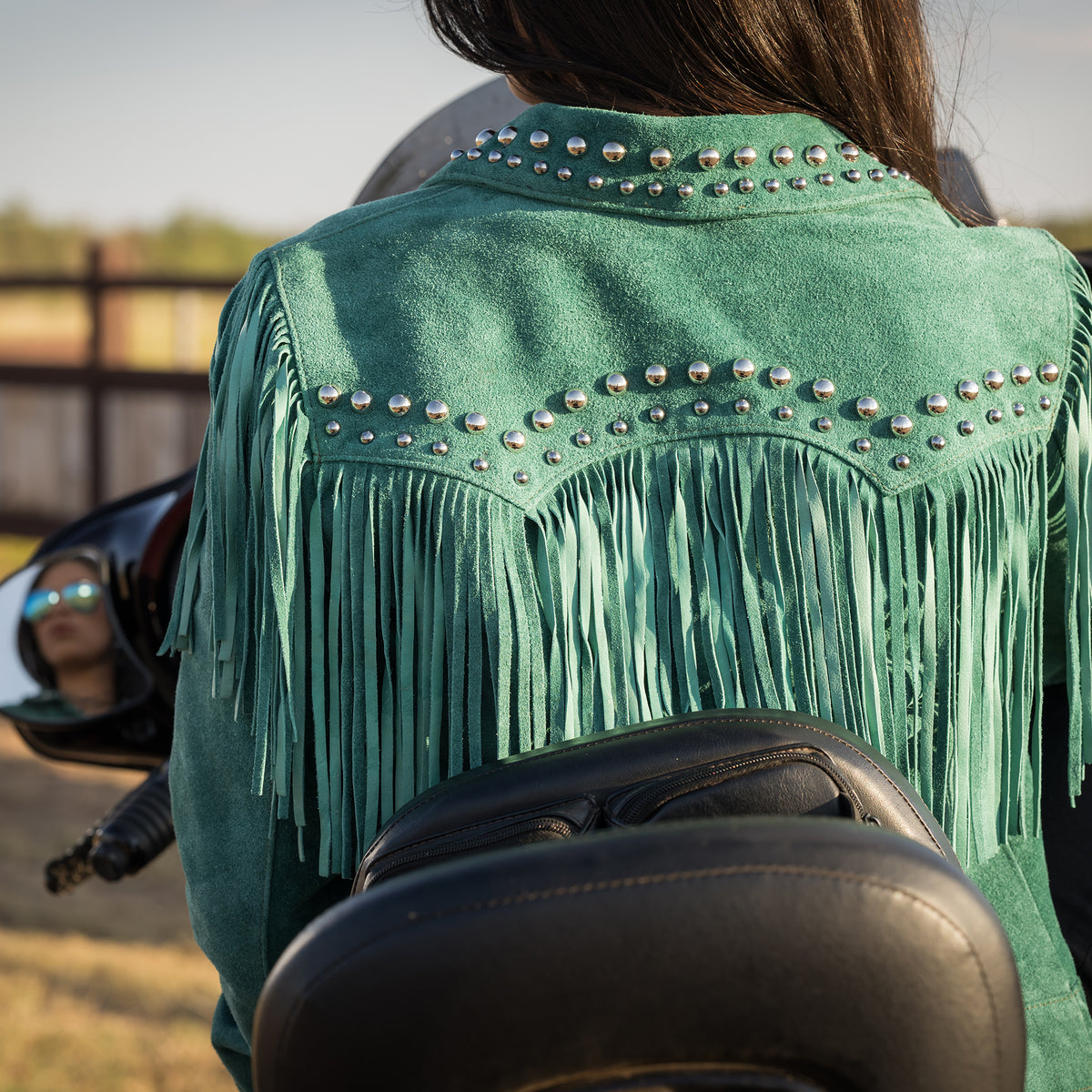 Closeup of the back of Soni Clifford on a black motorcycle wearing our emerald green suede jacket with hand studded detailing, fringe and snap front closure.