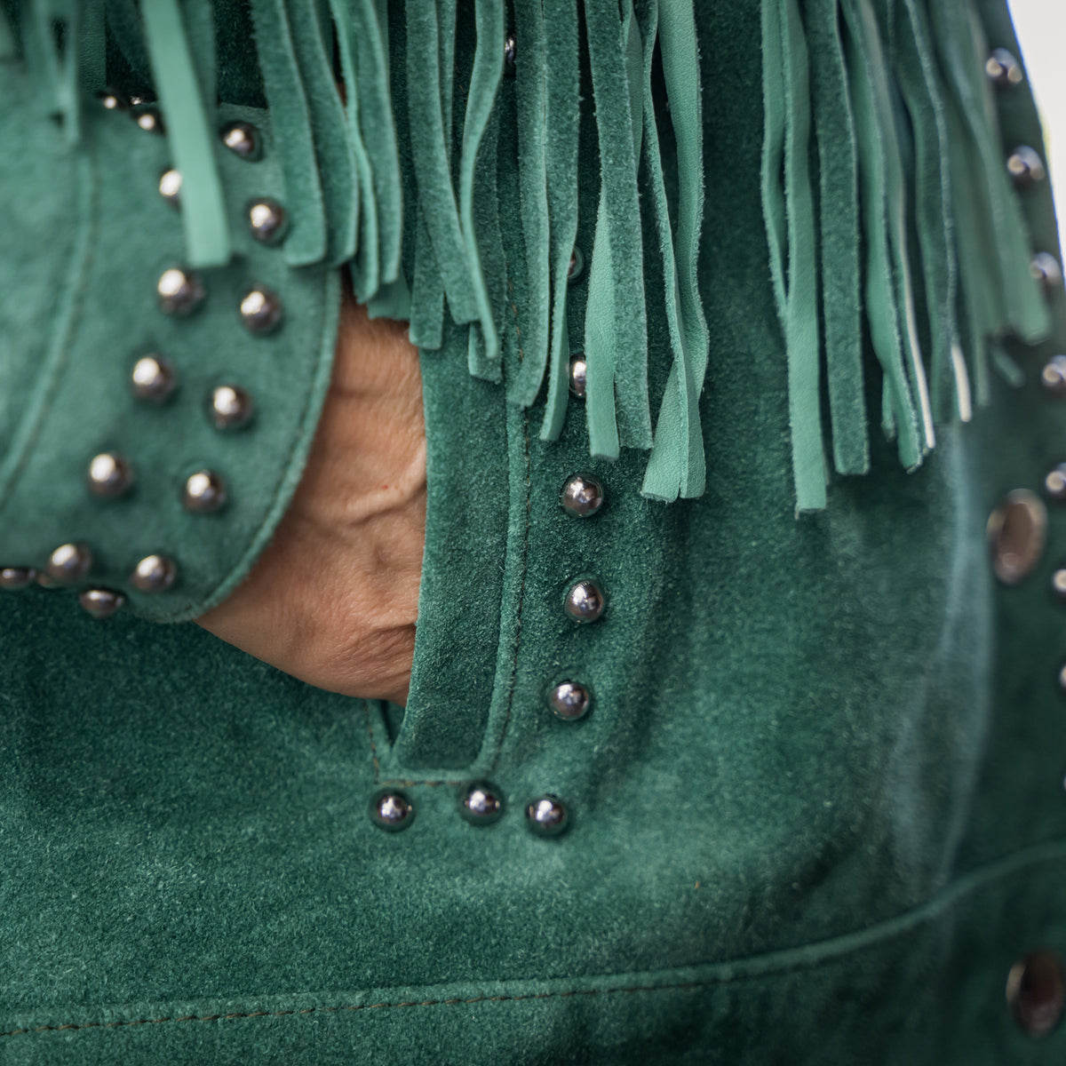 Closeup of a hand in the pocket of our emerald green suede jacket with hand studded detailing, fringe and snap front closure.