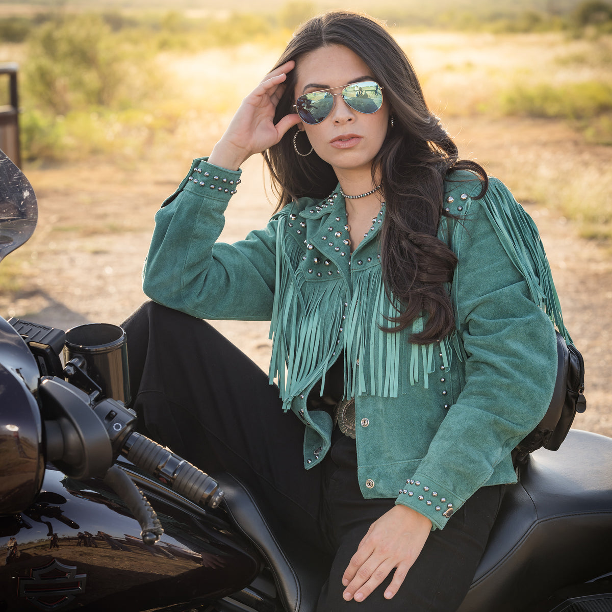 Soni Clifford on a black motorcycle wearing our emerald green suede jacket with hand studded detailing, fringe and snap front closure.