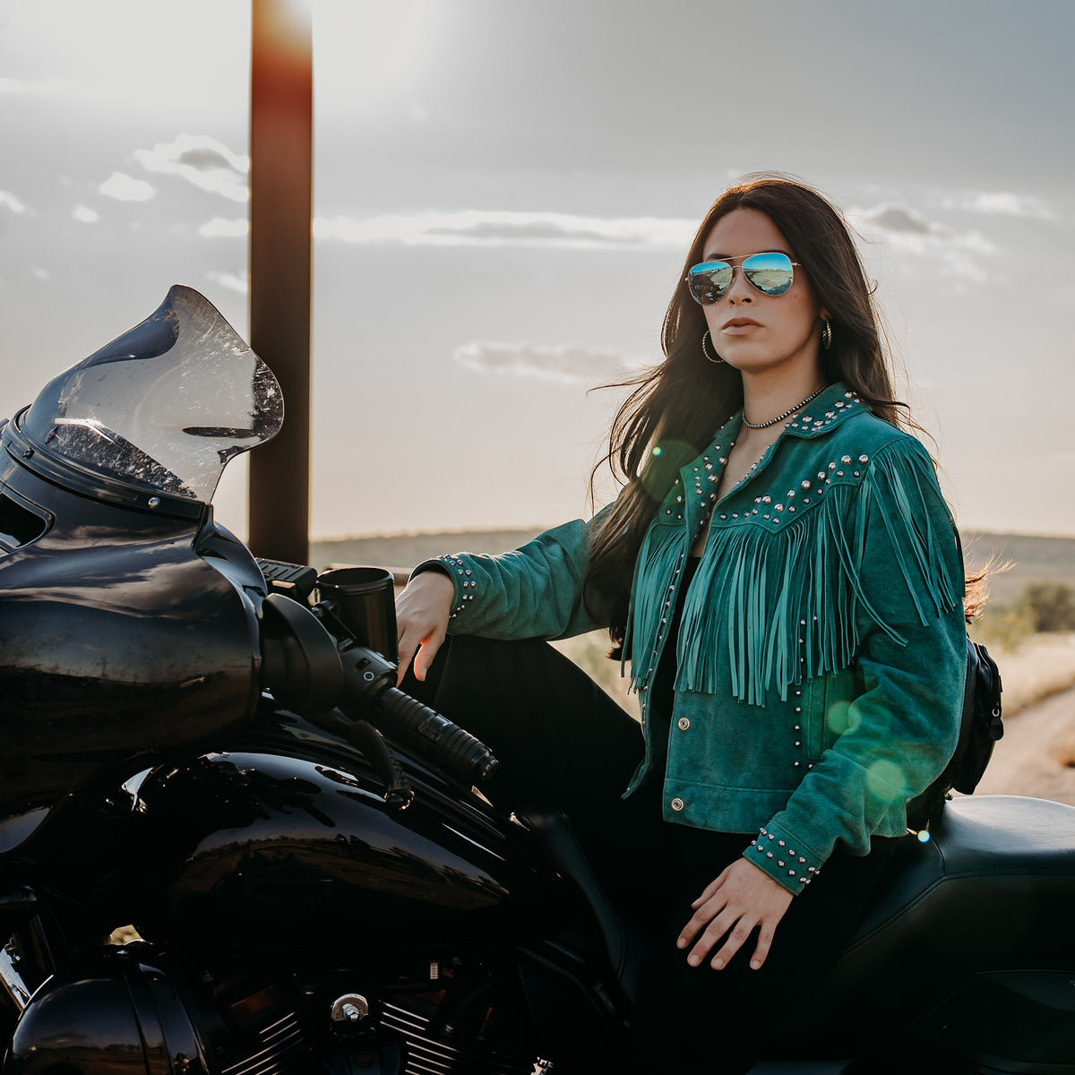 Soni Clifford on a black motorcycle wearing our emerald green suede jacket with hand studded detailing, fringe and snap front closure.