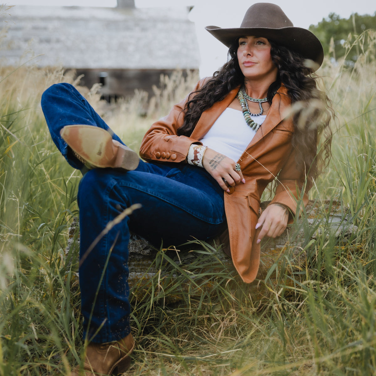 A woman sits in tall grass outdoors wearing a brown wide-brim hat, tan leather jacket, blue jeans, white top, and brown boots, with layered necklaces and bracelets.