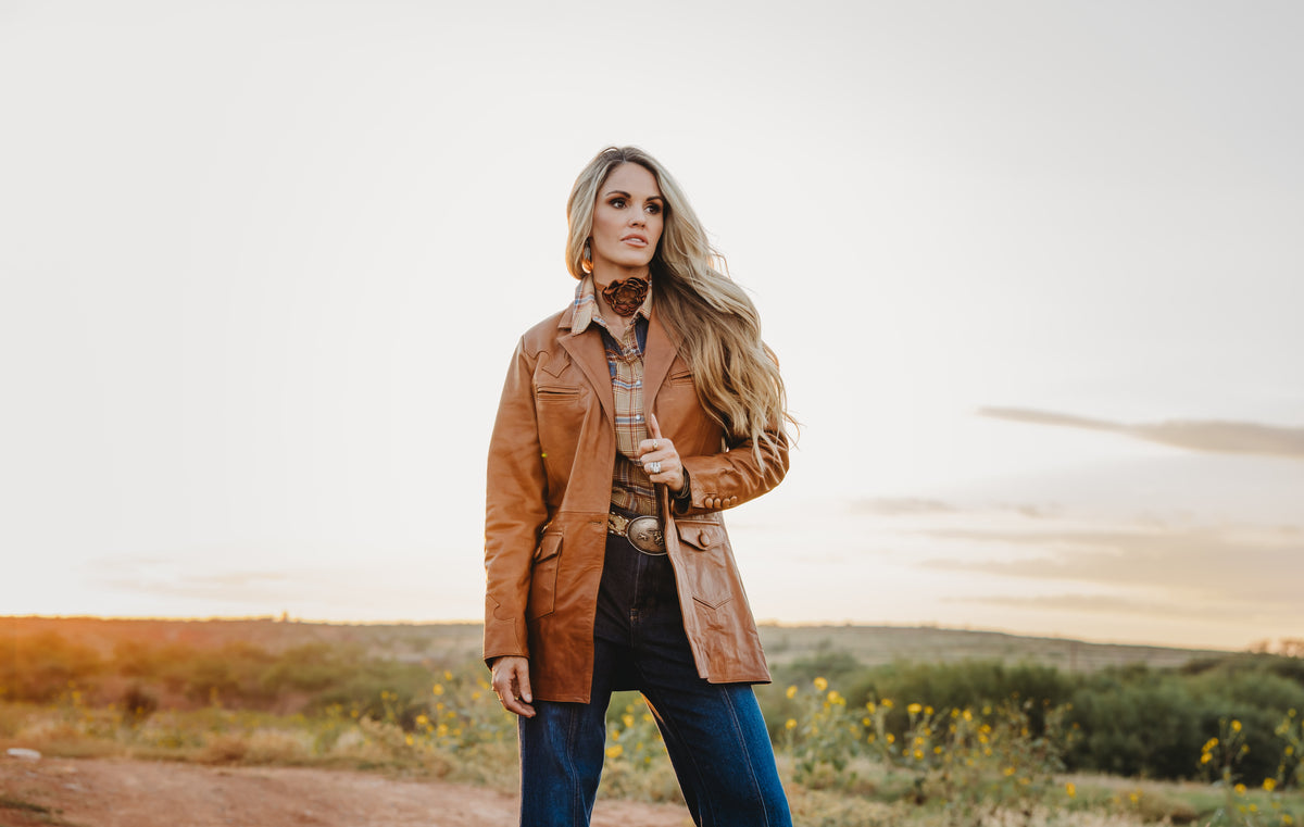 A woman stands outdoors in a grassy field at sunset wearing a brown leather jacket over a plaid shirt and blue jeans.