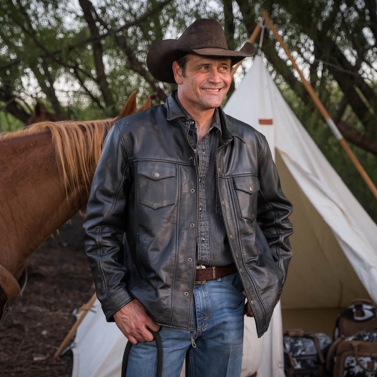 Stran T Smith wearing a brown cowboy hat, jeans and a men's black genuine leather rancher jacket with long sleeves, chest pockets and snap front closure.