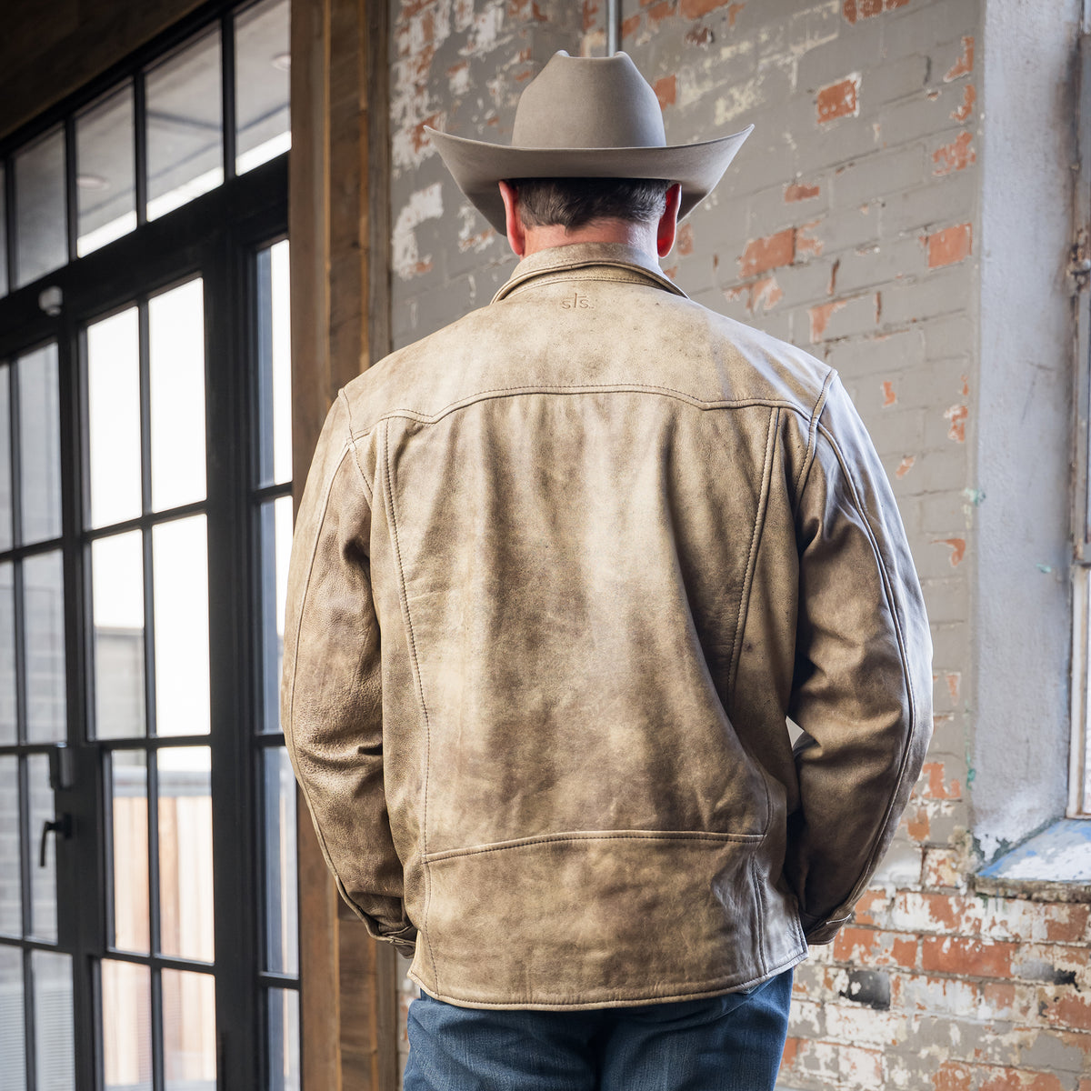 back of Stran Smith wearing our dappled dun beige leather jacket with front snap closure, two chest pockets, and hand warming pockets in front of a brick wall