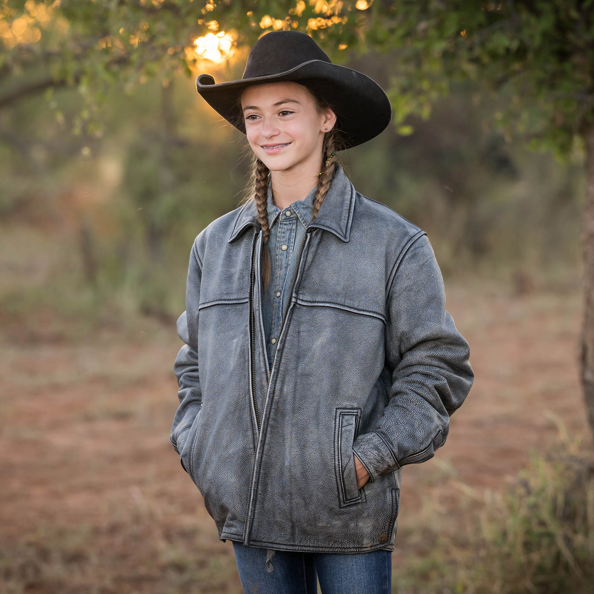 Selah Smith wearing jeans, black cowboy hat and stonewash genuine leather Rifleman jacket standing in the woods.