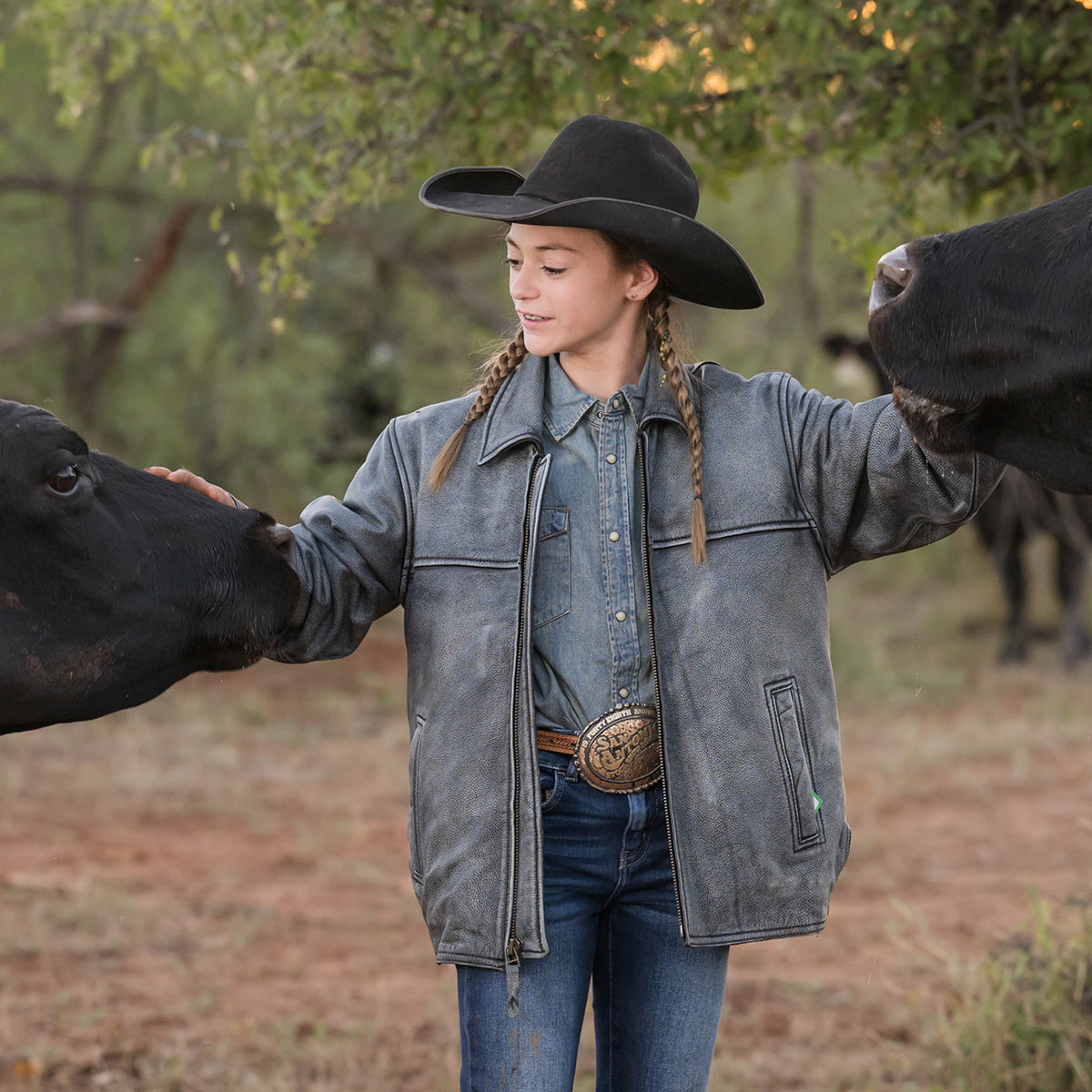 Selah Smith wearing jeans, black cowboy hat and stonewash genuine leather Rifleman jacket petting black angus cows.