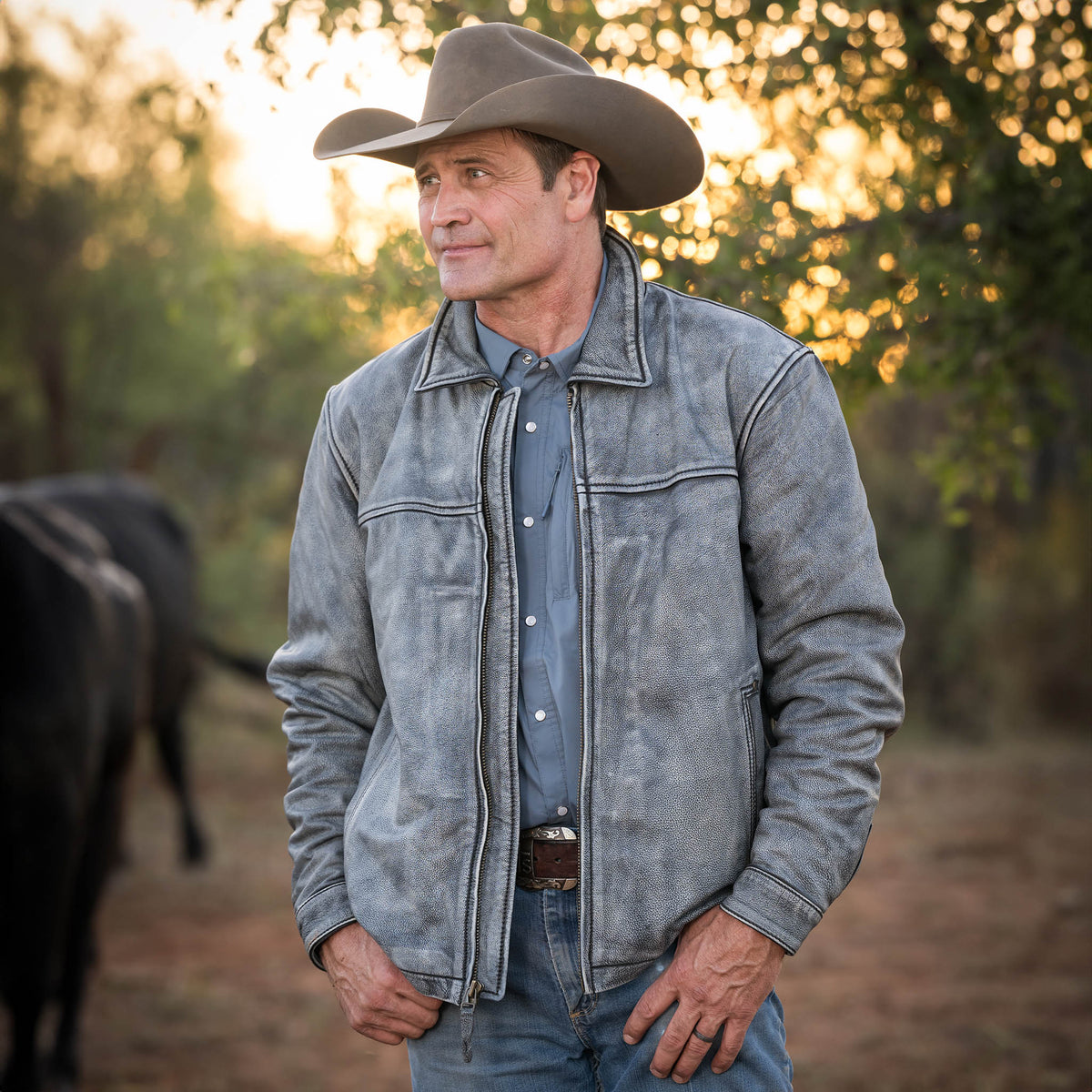 Stran T Smith wearing a tan cowboy hat, blue jeans and stonewashed genuine leather Rifleman jacket standing in the woods.