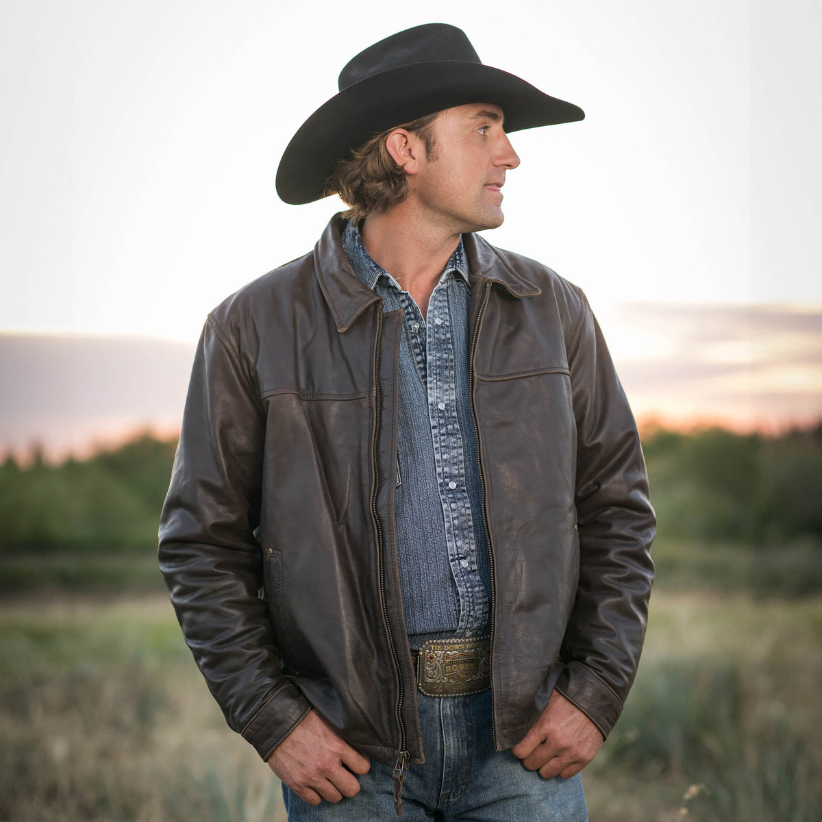 Man standing in a field wearing a black cowboy hat, jeans, denim shirt and brown genuine leather Rifleman jacket.