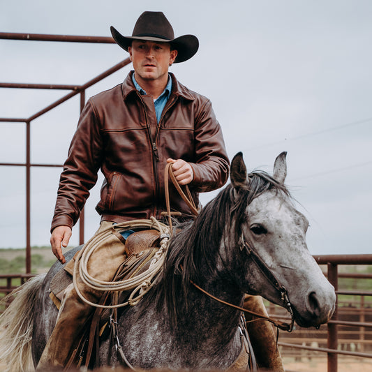 A man wearing a black cowboy hat and brown leather jacket rides a gray horse in an outdoor ranch setting with metal fencing visible in the background.