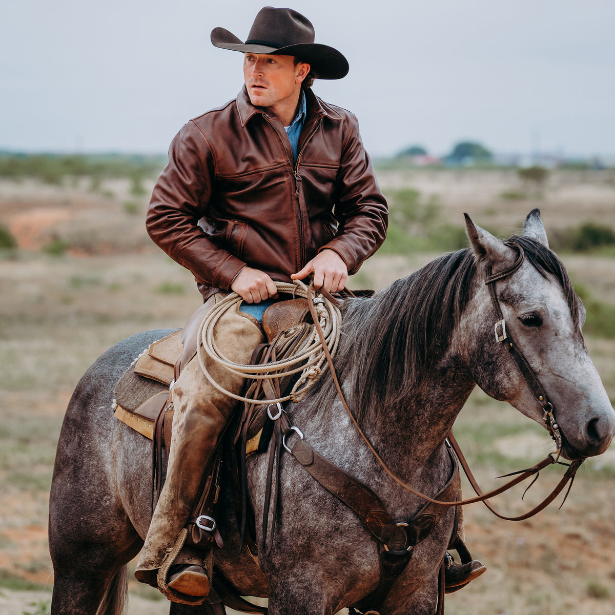 A man wearing a brown leather jacket and cowboy hat rides a dappled gray horse across an open outdoor landscape, holding a coiled lasso.