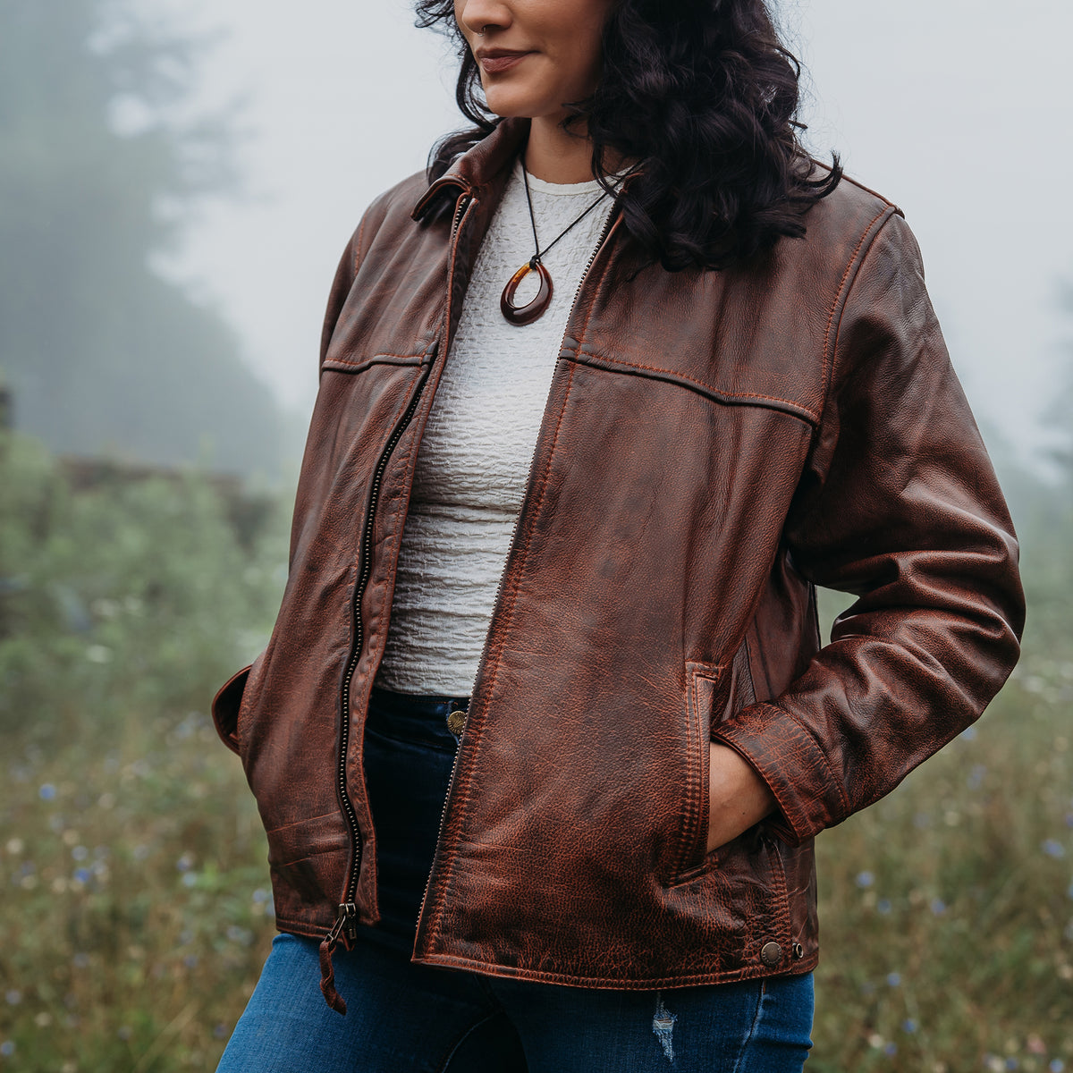 A woman wearing a brown leather jacket over a white top and blue jeans, standing outdoors in a misty grassy field.