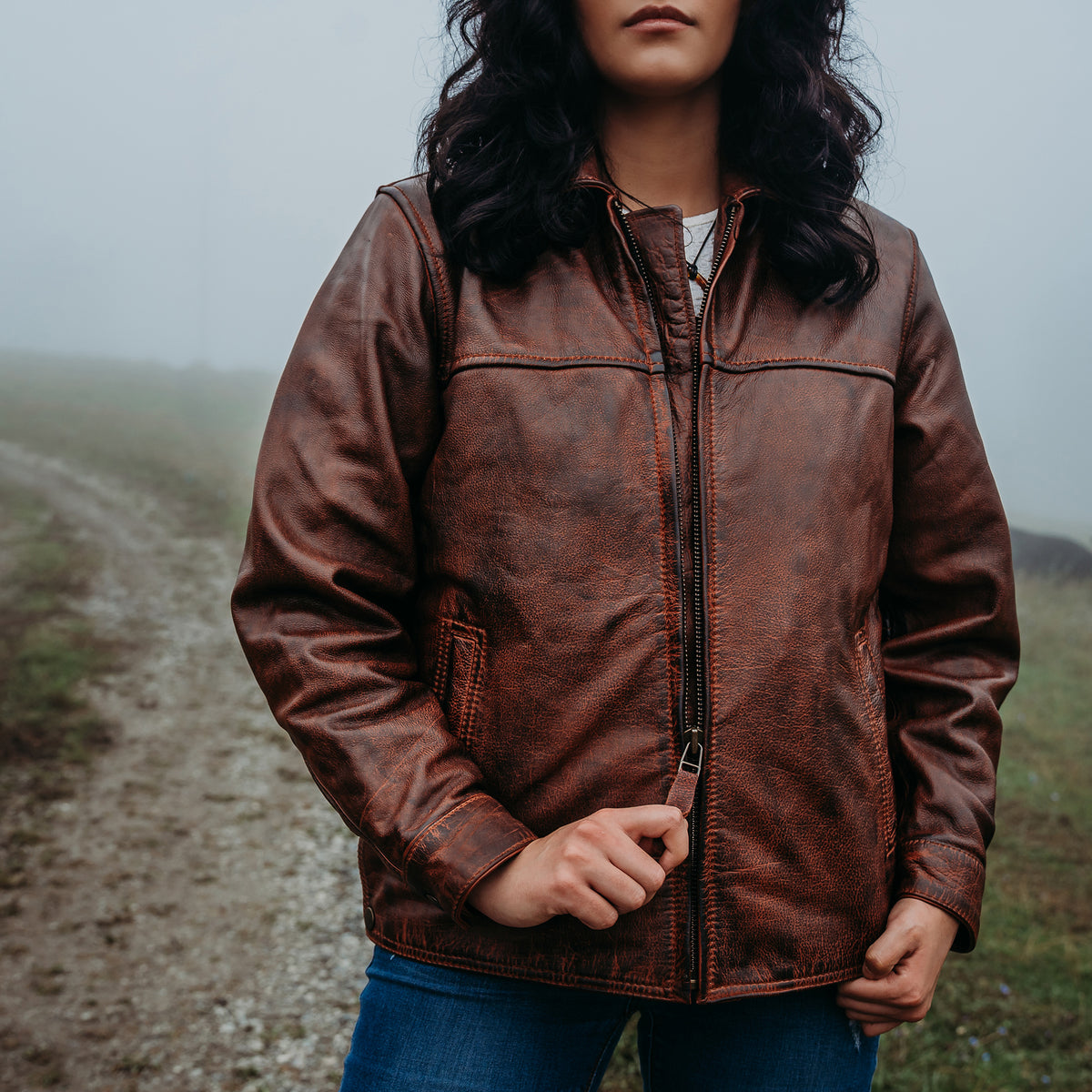A woman wearing a distressed brown leather jacket with a front zipper, standing on a misty dirt path outdoors.