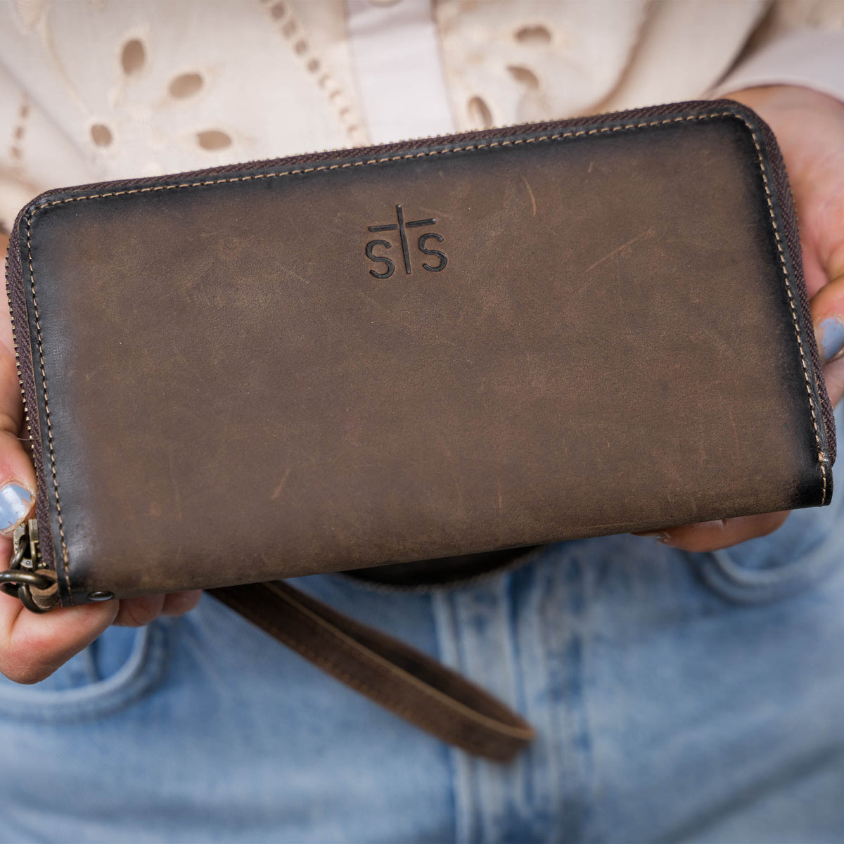 Closeup of a woman in a beige shirt holding an STS embossed leather wristlet with zip around closure.