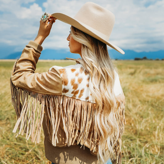 A woman in a cowboy hat and tan fringe jacket stands in a grassy field, seen from behind and to the side, looking up toward the sky.