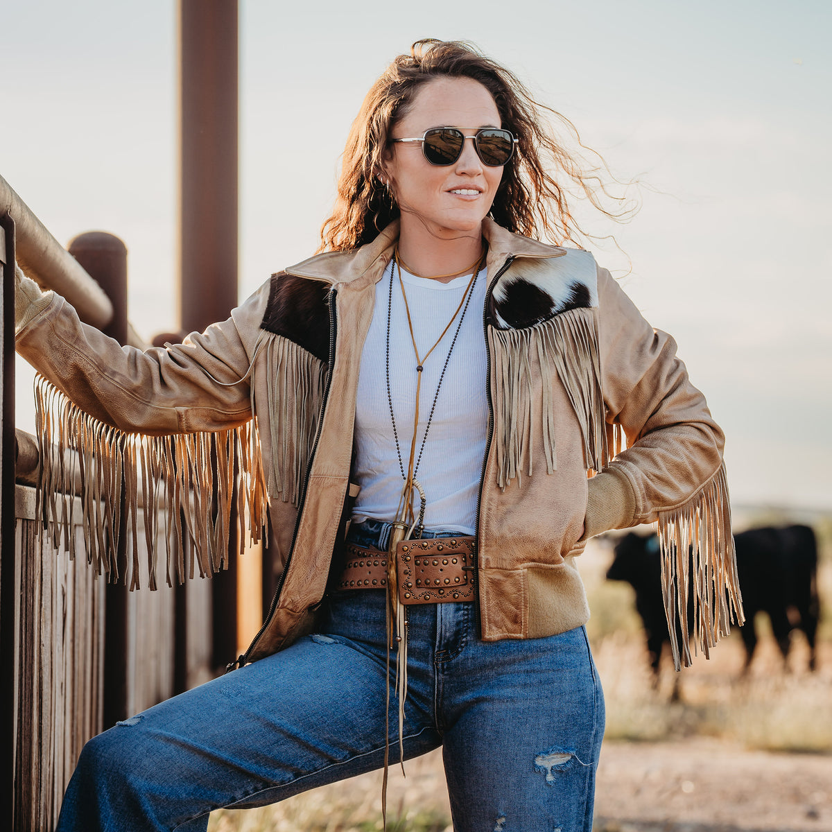 Woman in sunglasses wearing our beige leather jacket with cowhide detailing, fringe and zip front closure outside in front of cattle.