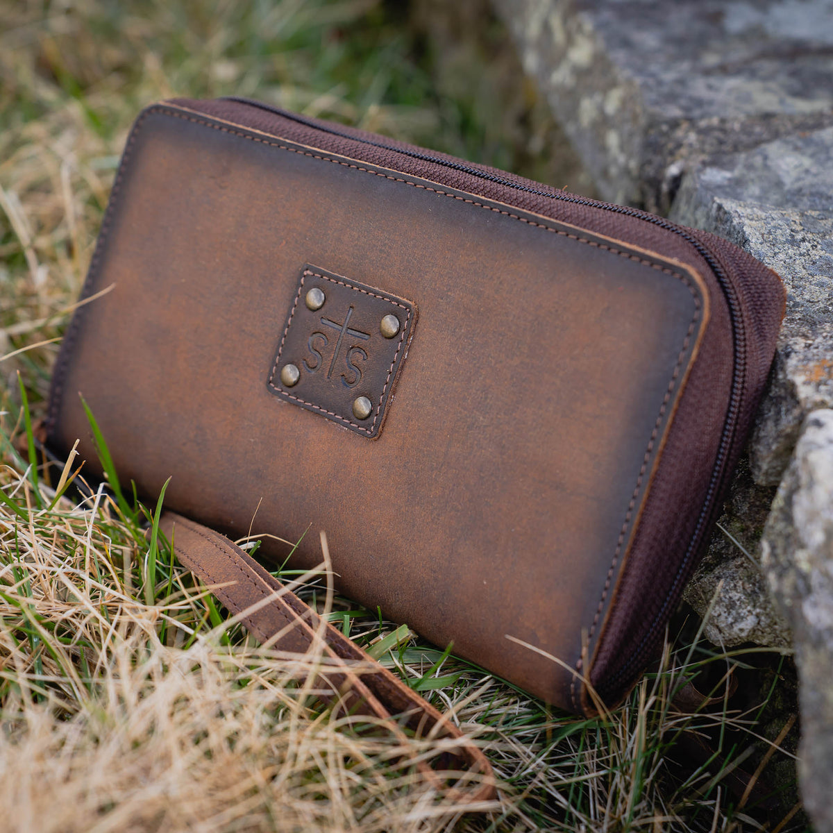 Brown genuine leather wallet with zip around closure, leather STS branded patch and leather wristlet strap laying in the grass.
