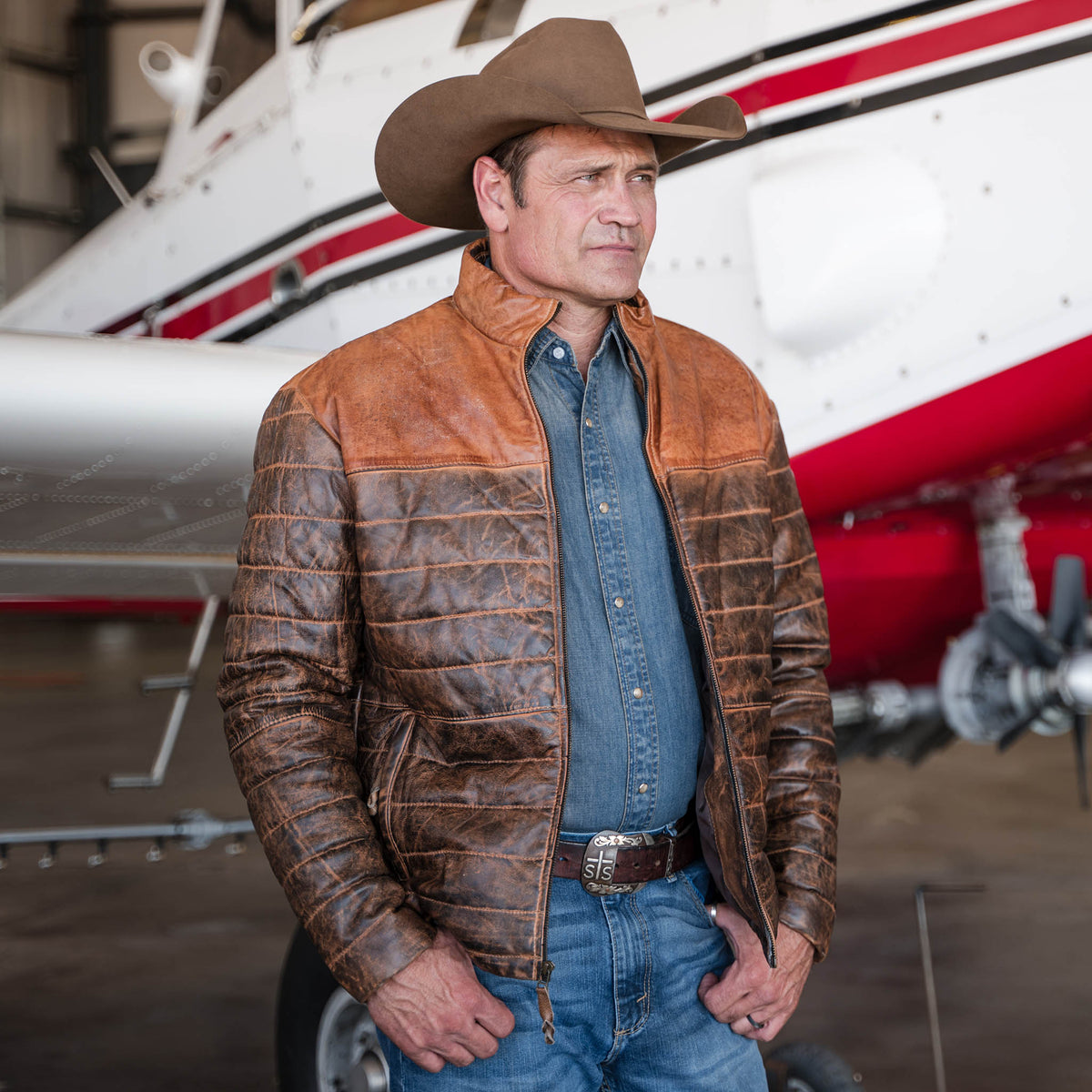 Stran T Smith wearing a tan cowboy hat, denim shirt, jeans and a brown men's Clyde leather puffer jacket with a full-grain leather exterior standing in front of a small airplane.