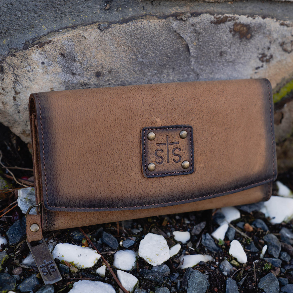 Brown leather trifold wallet with leather flap closure and zipper closure and "STS" stamped on a leather patch sitting on a rock.