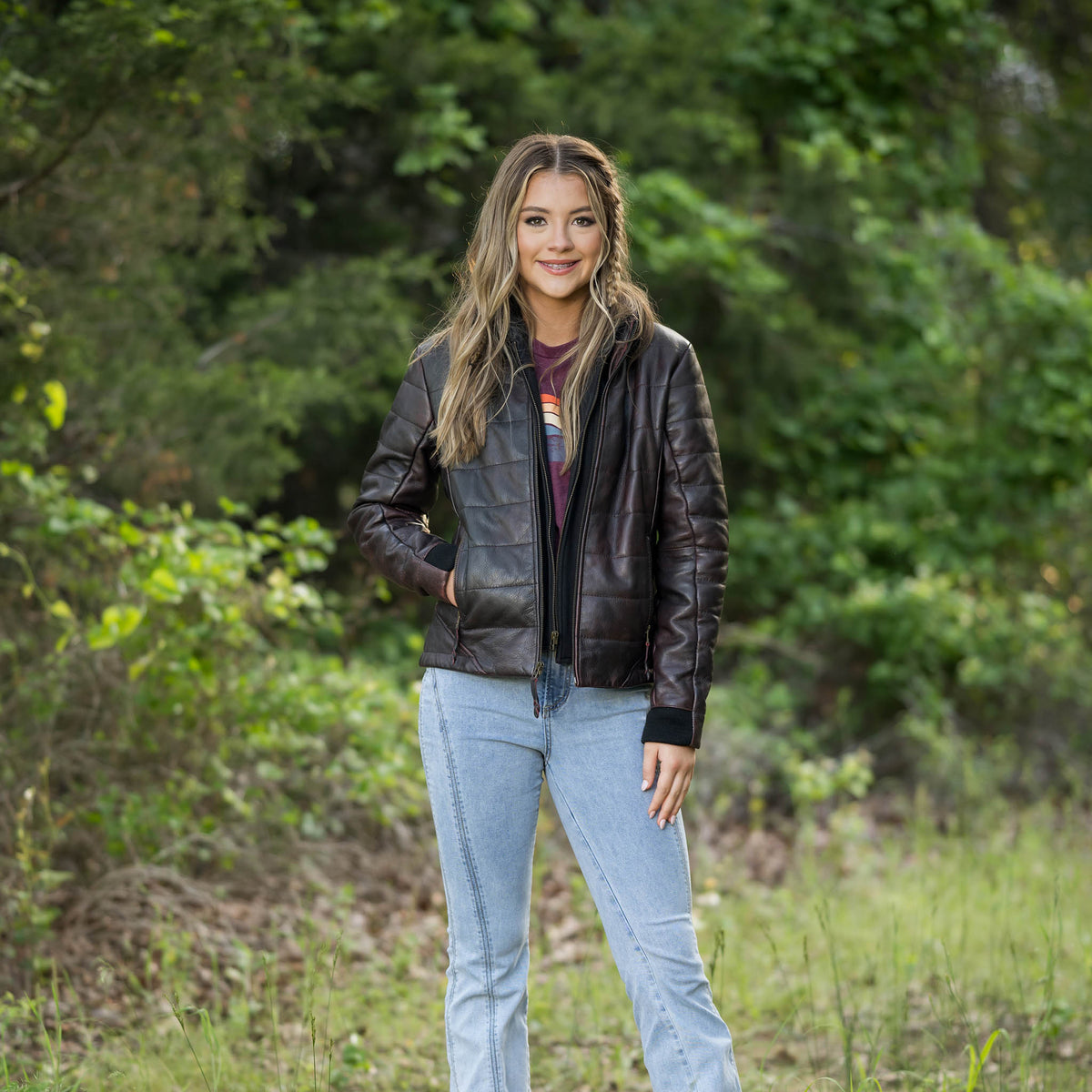 Woman wearing a black leather jacket and light blue jeans standing in a forested area.