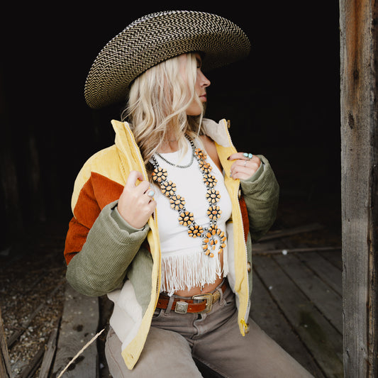 A woman wearing a wide-brim woven hat, a white fringed top, a yellow and olive green color-block jacket, and brown pants, seated in a rustic wooden doorway.