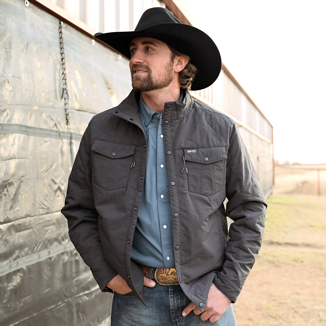 Man wearing a black cowboy hat, jeans and a men's lightweight quilted poly field jacket with snap front closure, multiple exterior pockets, and snap cuffs on a ranch setting.